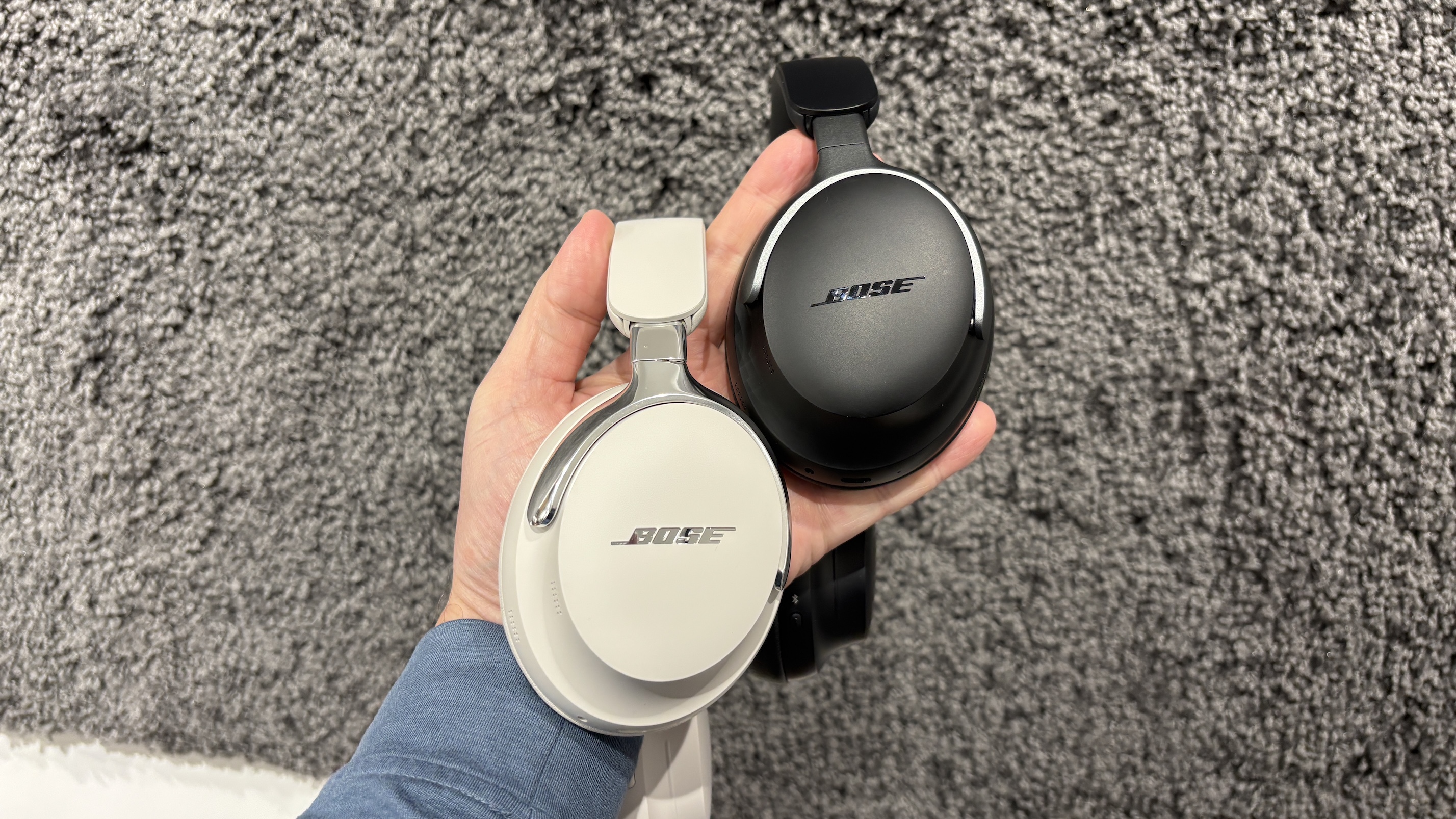 A pair of Bose QuietComfort Ultra Headphones (2nd Gen) and their first-gen predecessors being held in one hand in front of a grey fluffy surface.