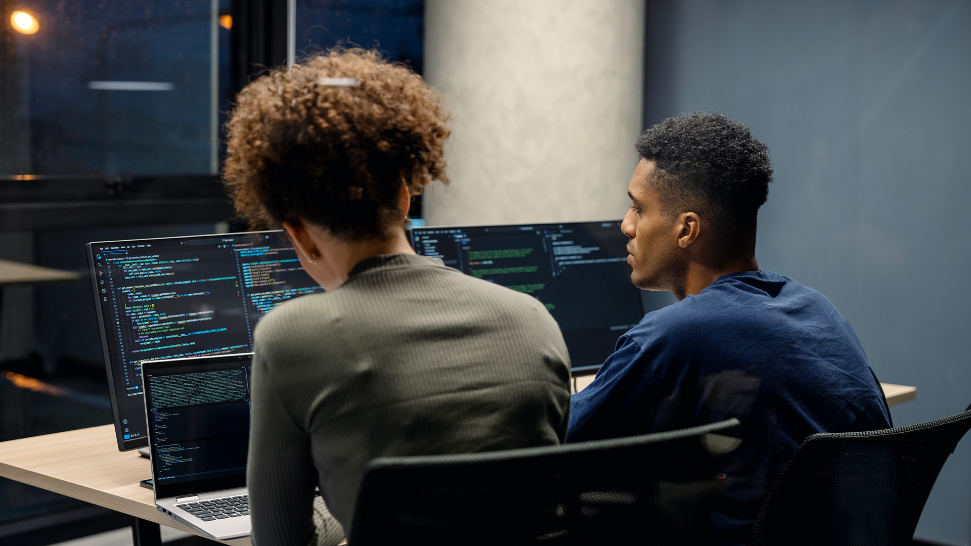 Male and female software developers sitting side by side coding on desktop computers, with computer code pictured on monitors.