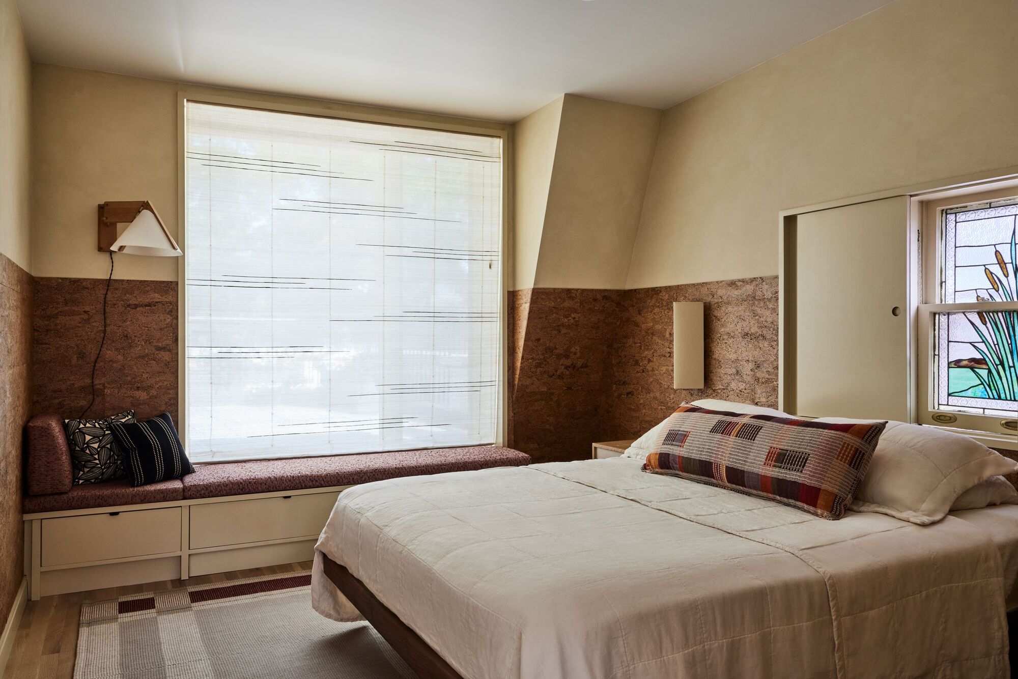 bedroom with window seat, blinds, cork wall wrap, and plaster ceiling, mirror behind the bed, and cream bedding with patterned cushion