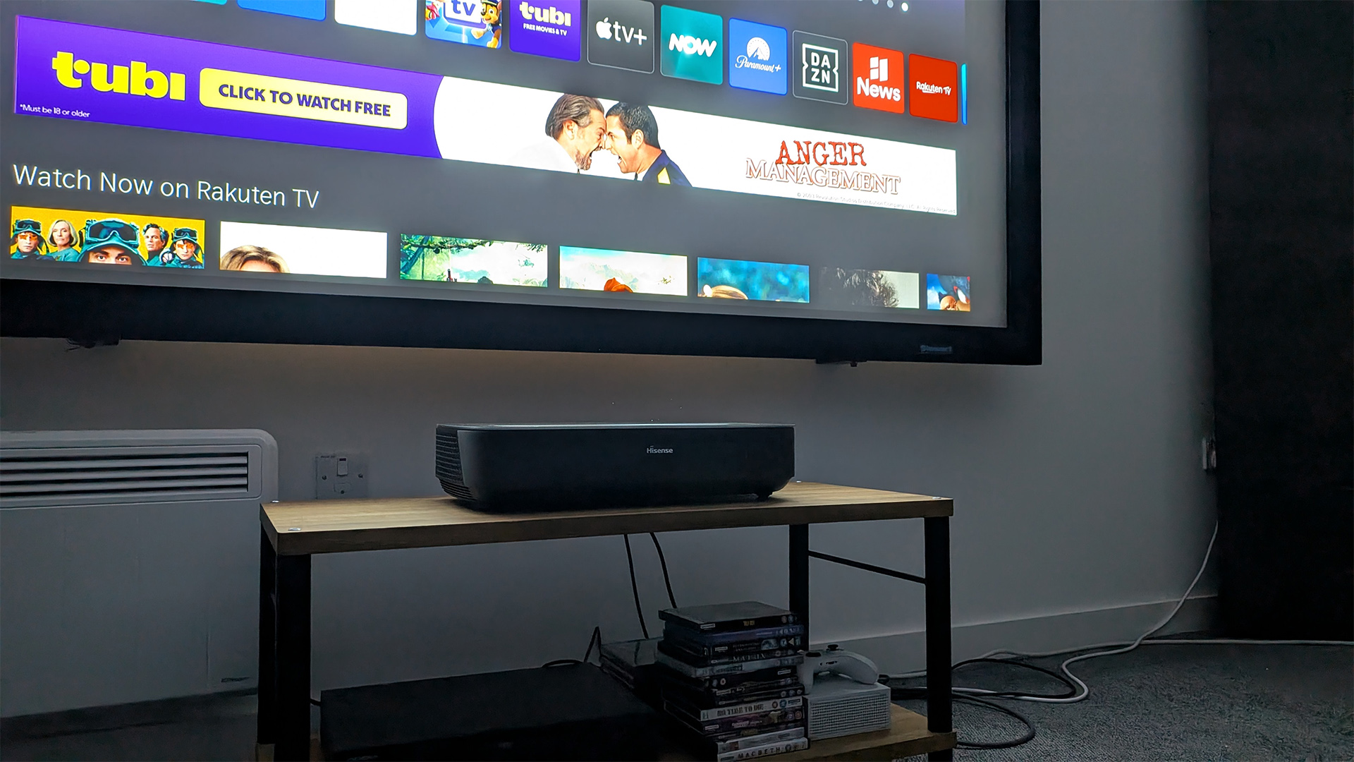 Hisense PL2 UST projector on wooden AV rack in test room. On screen is VIDAA OS home screen