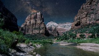 a river flows toward the foreground of the image with large rock structures behind and a star studded sky in the distance.