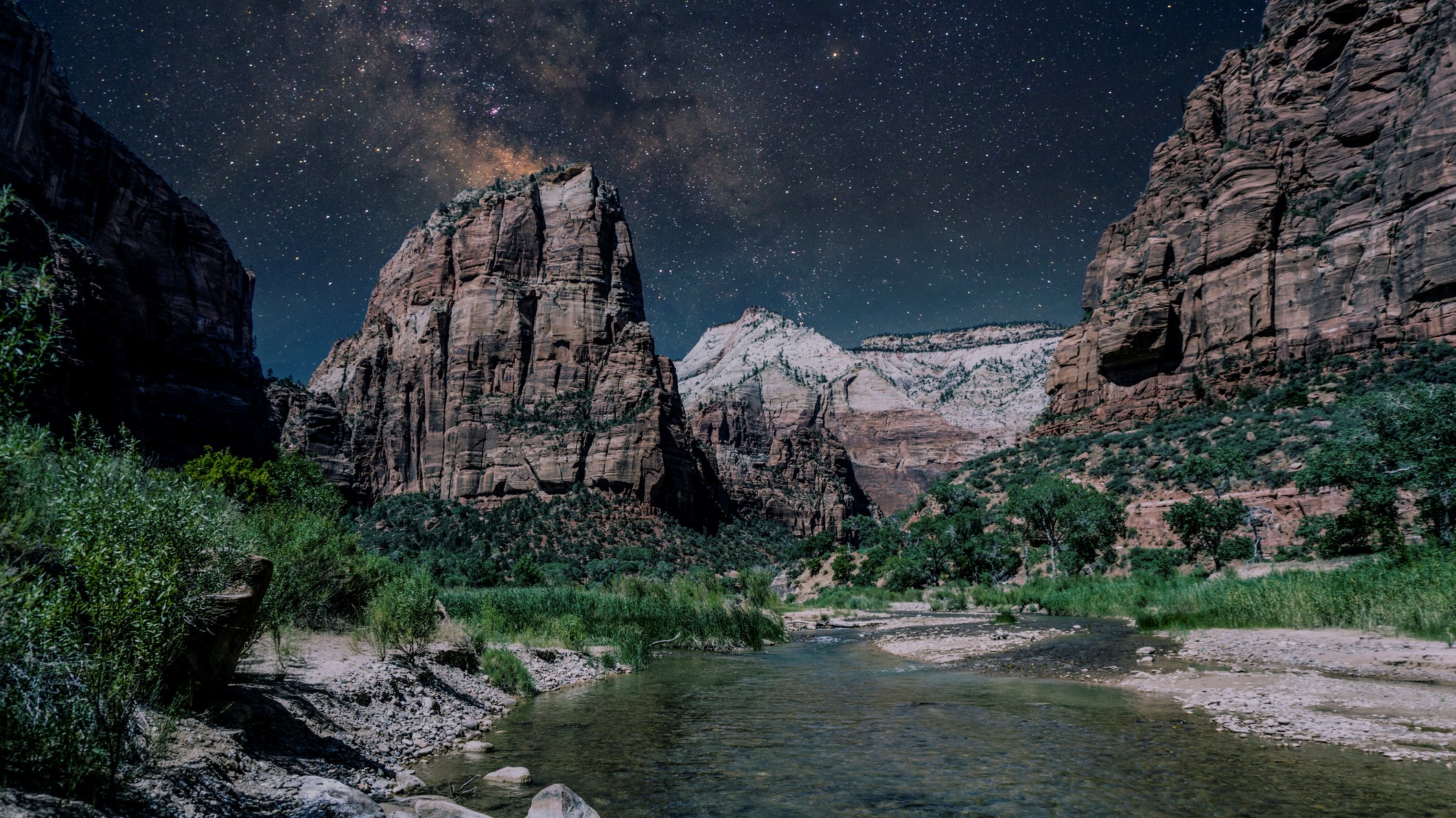 a river flows toward the foreground of the image with large rock structures behind and a star studded sky in the distance.