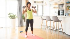 woman wearing a yellow vest and black leggings standing in her socks on a pink exercise mat in home setting. there's a window behind her and a kitchen and breakfast bar to her right with three high chairs.