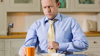 A businessman in a tie appears to have indigestion after eating. An empty plate sits in front of him.