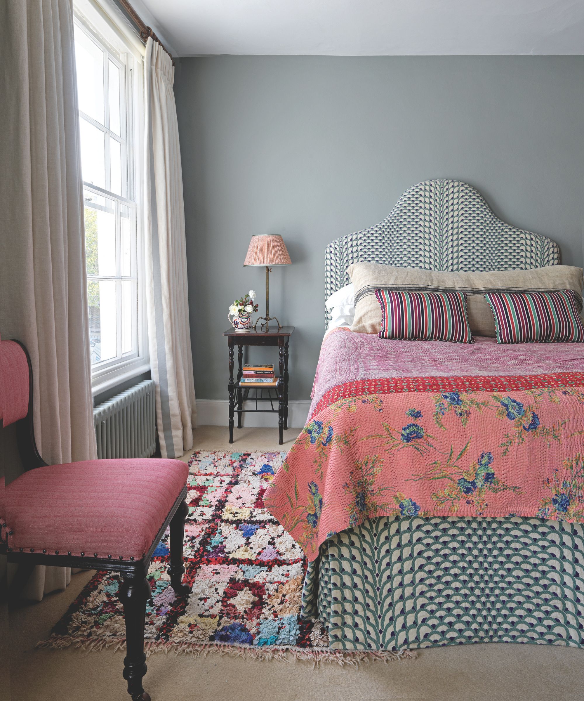 A sage green bedroom with a colorful patterned bed, colorful patterned rug and pink and dark wood upholstered chair.
