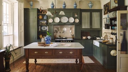 Cream kitchen with tall sunny window to the left, island dining table in the middle, green built-in cabinets and blue porcelain jars decorating the top of cabinets