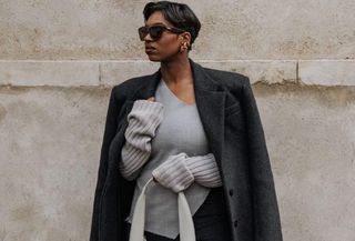 Influencer standing in front of wall wearing gray sweater, black blazer, and white Marge Sherwood bag.