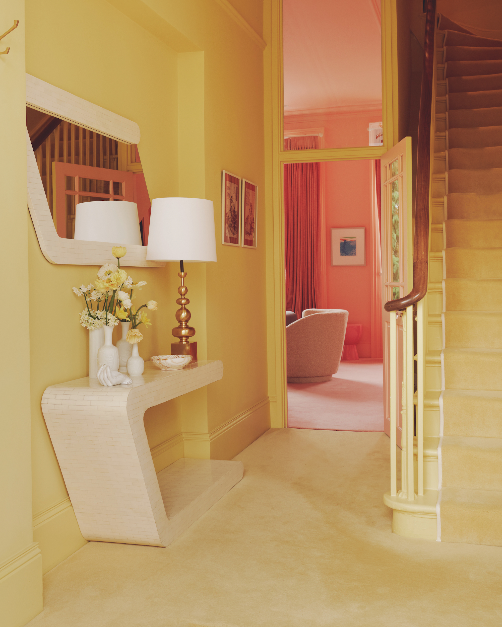 A butter yellow, color drenched entry way, with a view up the staircase and into a peachy pink, living room. On the felt hand wall in the entryway is a modern side table with a mirror above.