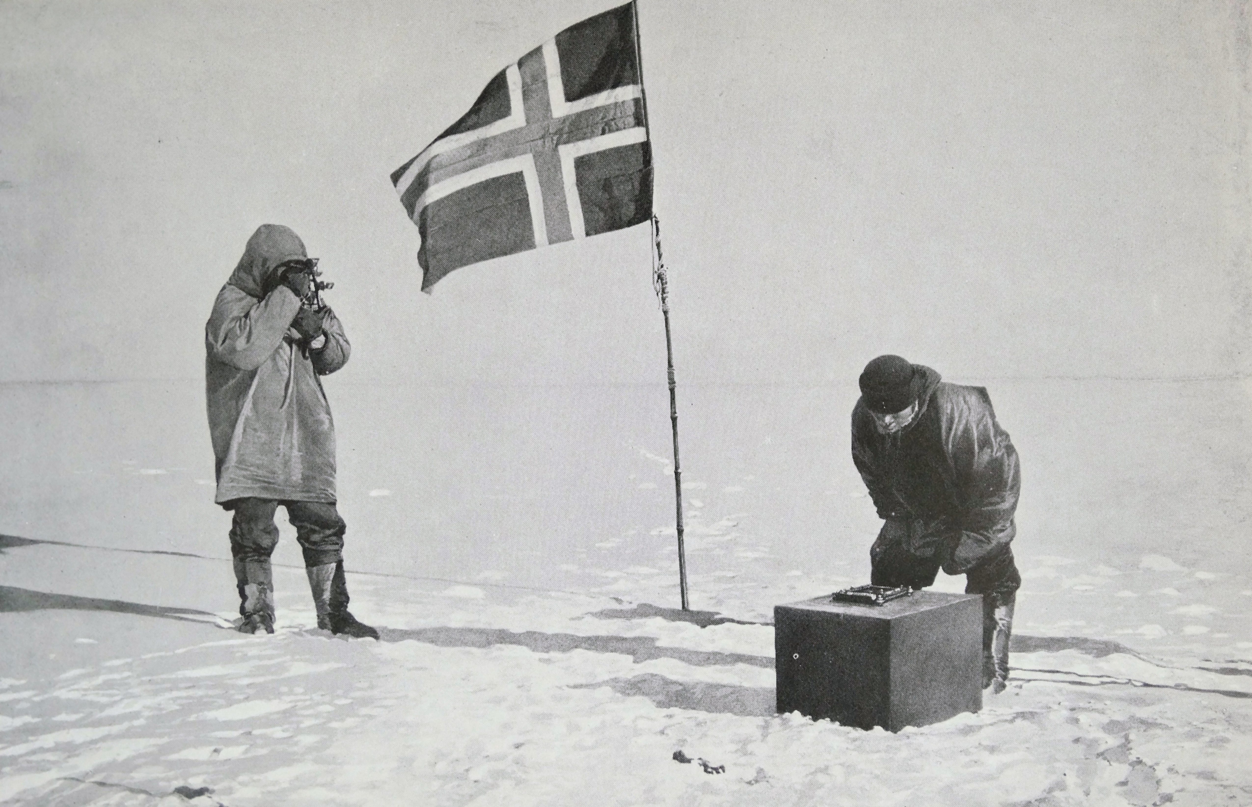 Black and white photo of Capt. Roald Amundsen standing and looking through binoculars. He's next to a Norwegian flag planted in the snow. A man next to him is looking at a crate on the ground.