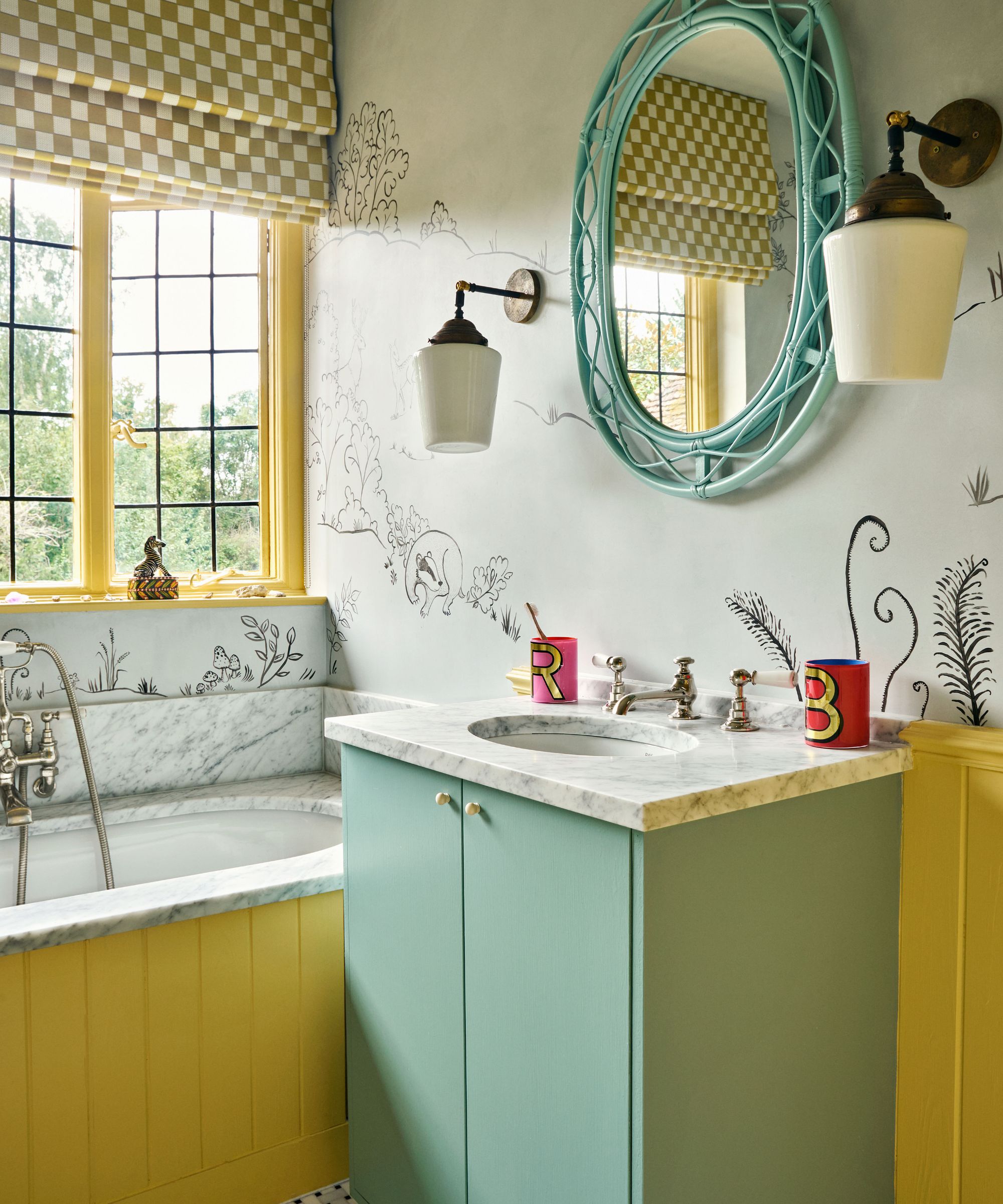 Bathroom with wallpapered walls, yellow wood panelling on the wall and bath, a vanity unit painted in sage with a marble sink, and a yellow window frame and chequerboard roman blind.