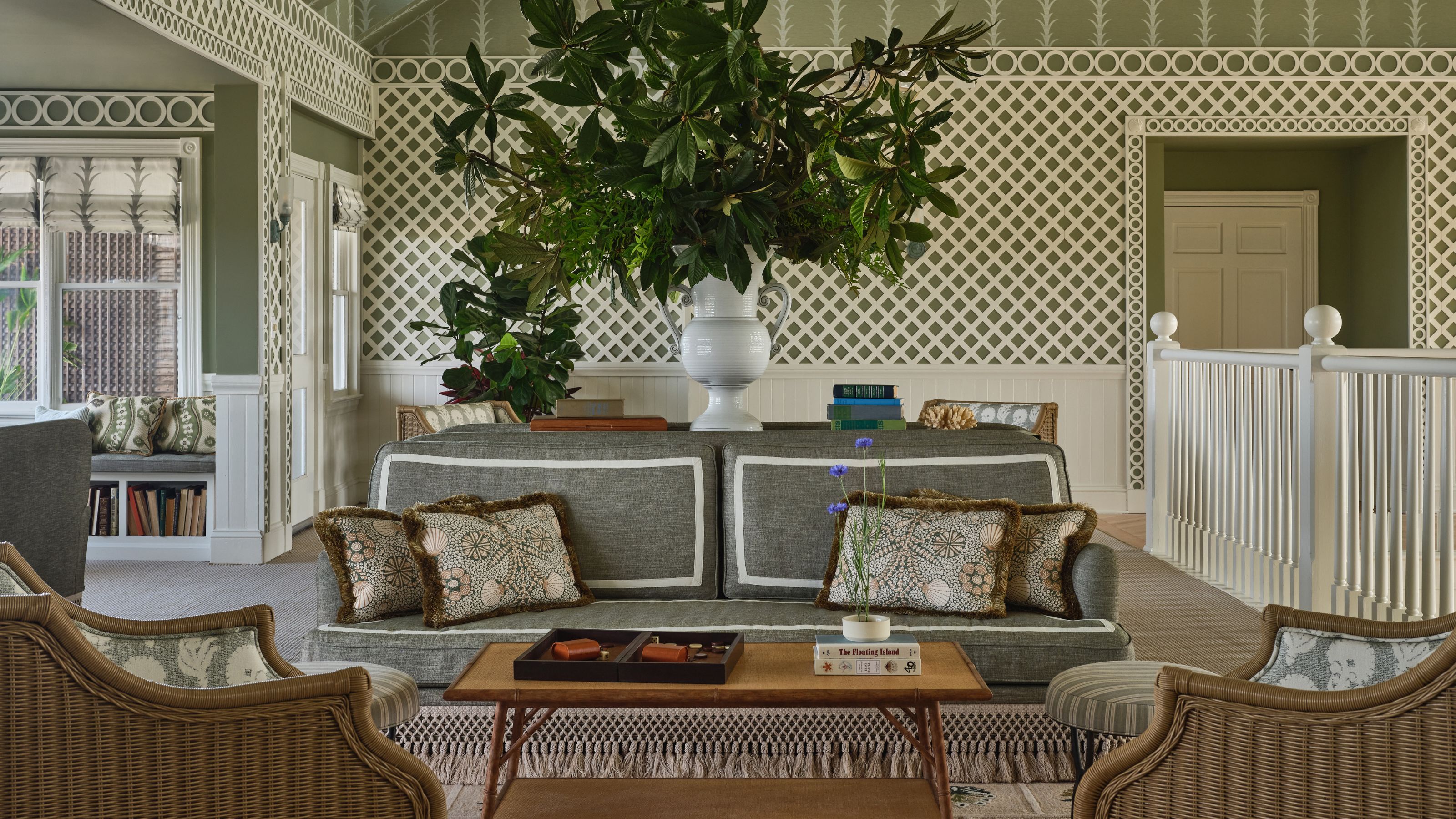 A sophisticated lounge area featuring a grey sofa with patterned pillows, wicker armchairs, white lattice-patterned walls, and a large central leafy plant arrangement.