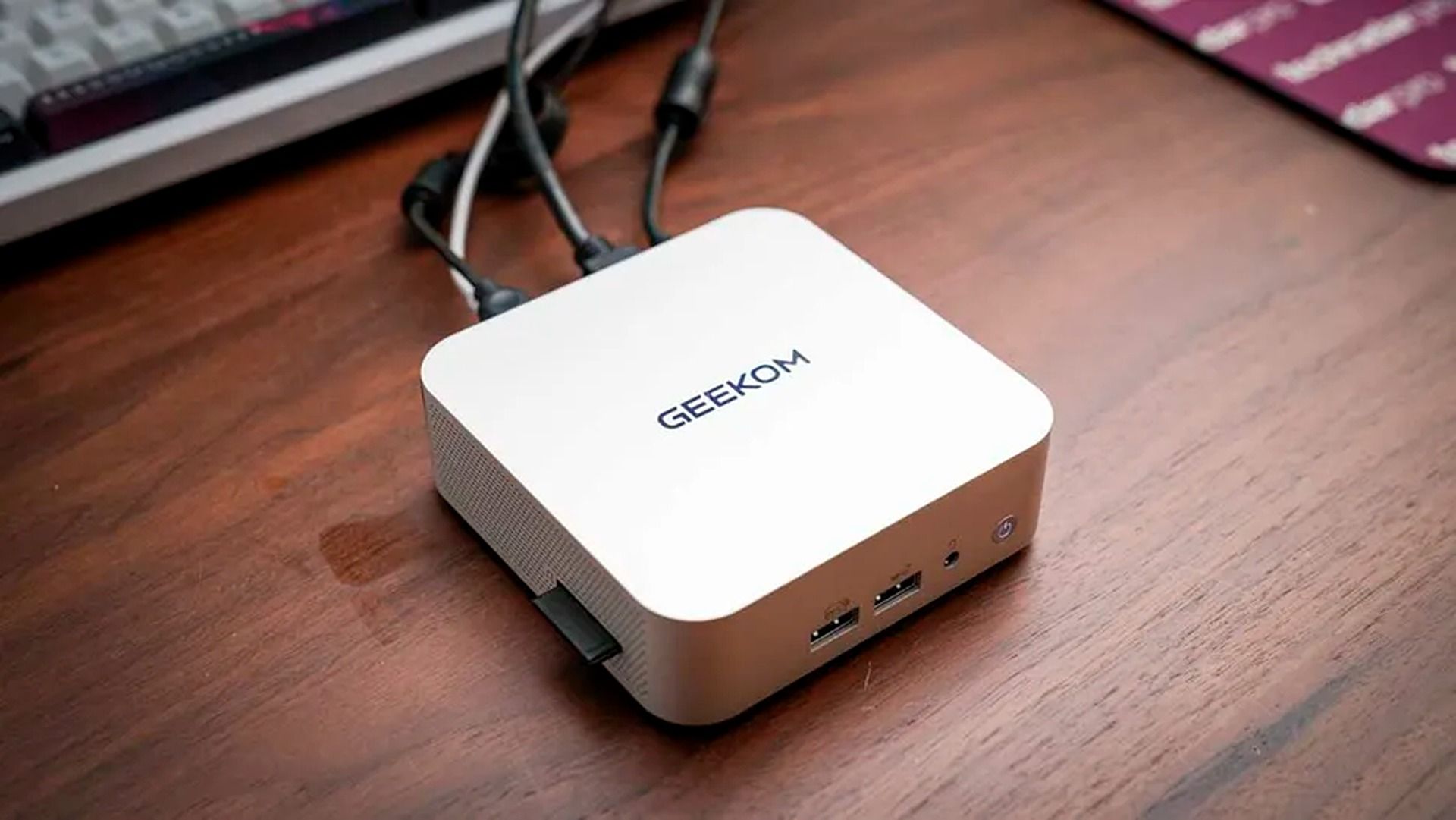 A compact white GEEKOM mini PC sits on a wooden desk, with cables connected and keyboard edge visible, creating a tidy, practical workspace.