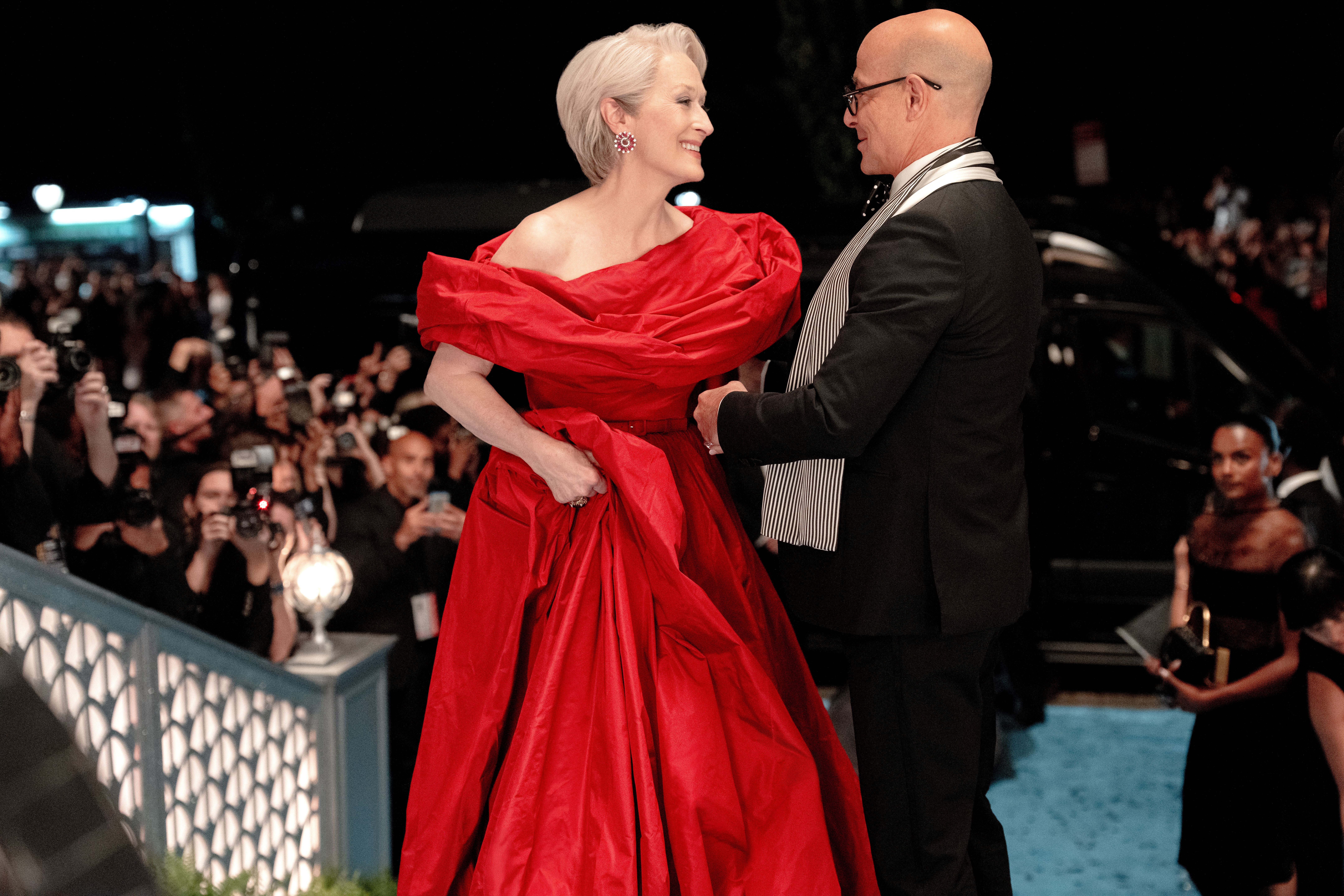 Meryl Streep as Miranda Priestly wearing a red gown talking to Stanley Tucci as Nigel Kipling in a black suit on a carpet as photographers take photos in the distance in the devil wears prada 2