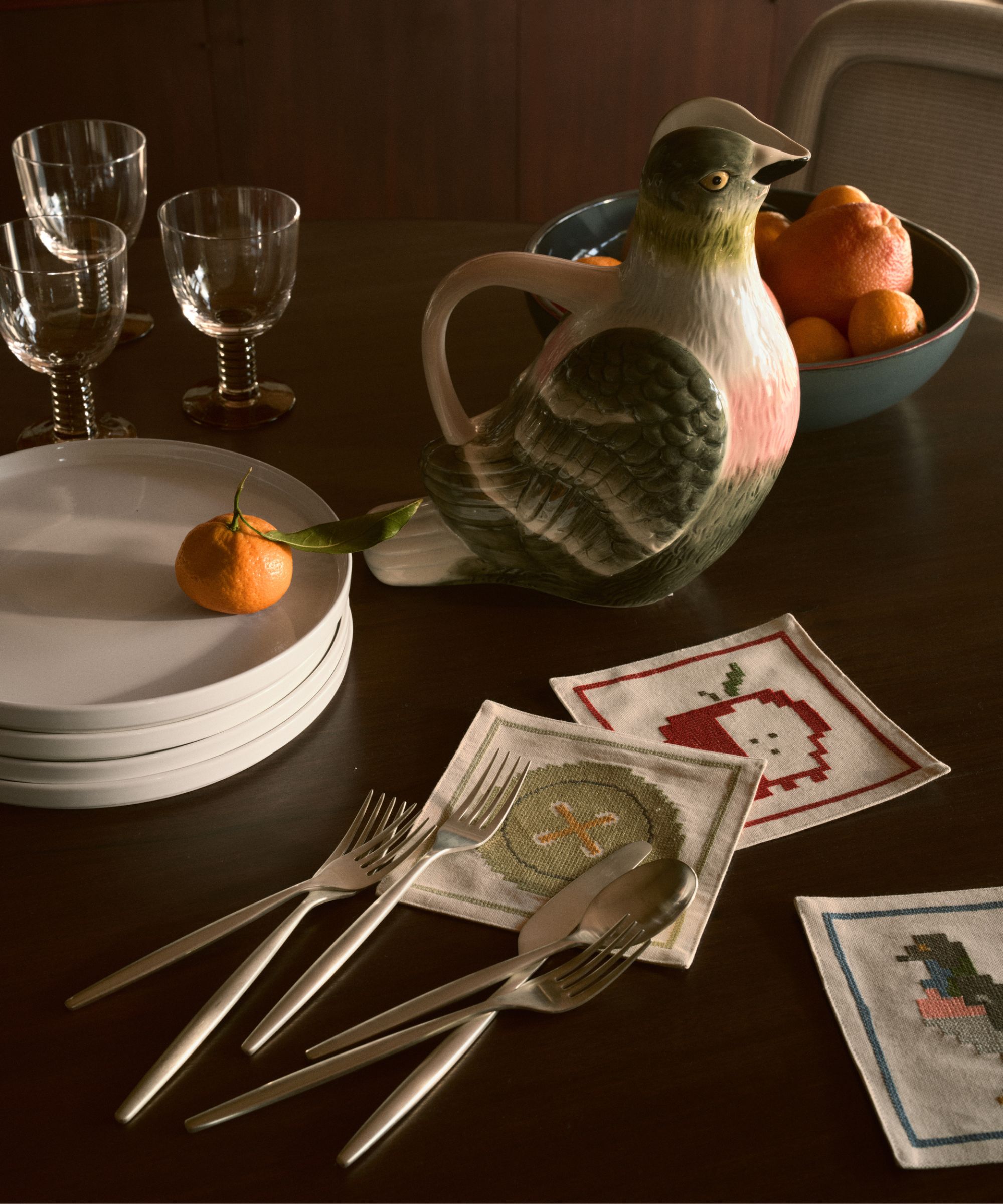 Image of a dark wooden table with a stack of white plates, a stack of silverware, embroidered cocktail napkins, a pigeon pitcher, and three wine glasses.