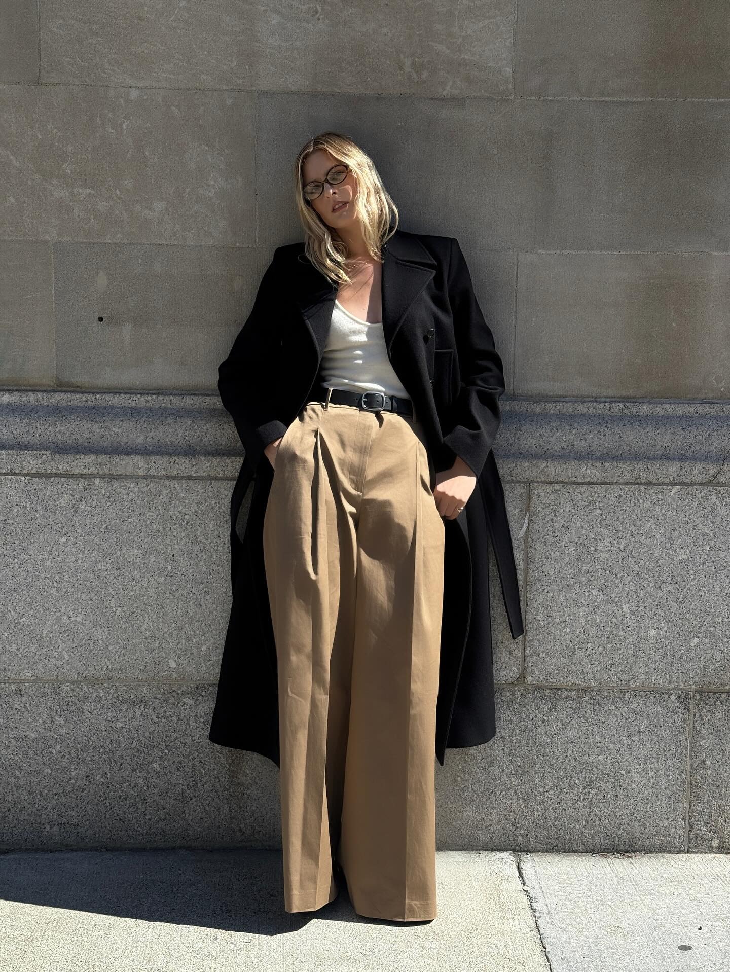 Christie wears a black long coat, white tank top, black belt, and khaki wide-leg trousers.