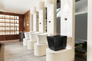Bathroom image, row of four intelligent toilet designs on individually raised stands by Kohler, light marble floor, dark wood wall to the far left with glass window display, silver sink, mirrors