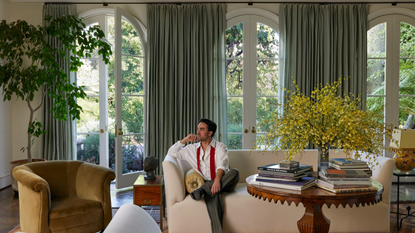 Jake Arnold posing on the cream sofa of a stately sunlit living room featuring a trio of tall arched windows dressed with powdery blue curtains