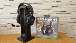 The SteelSeries Arctis Nova 5X gaming headset on a headset stand in front of a white brick background next to its box