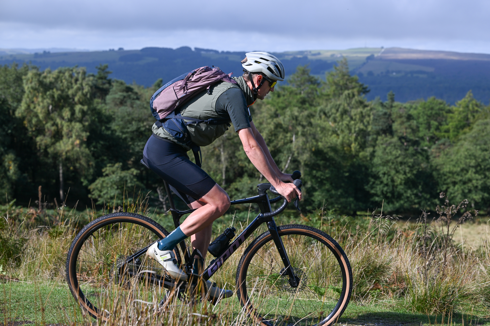 Man riding a gravel bike wearing a green jersey, black shorts and a green gilet with a pink and blue backpack on, viewed from the side
