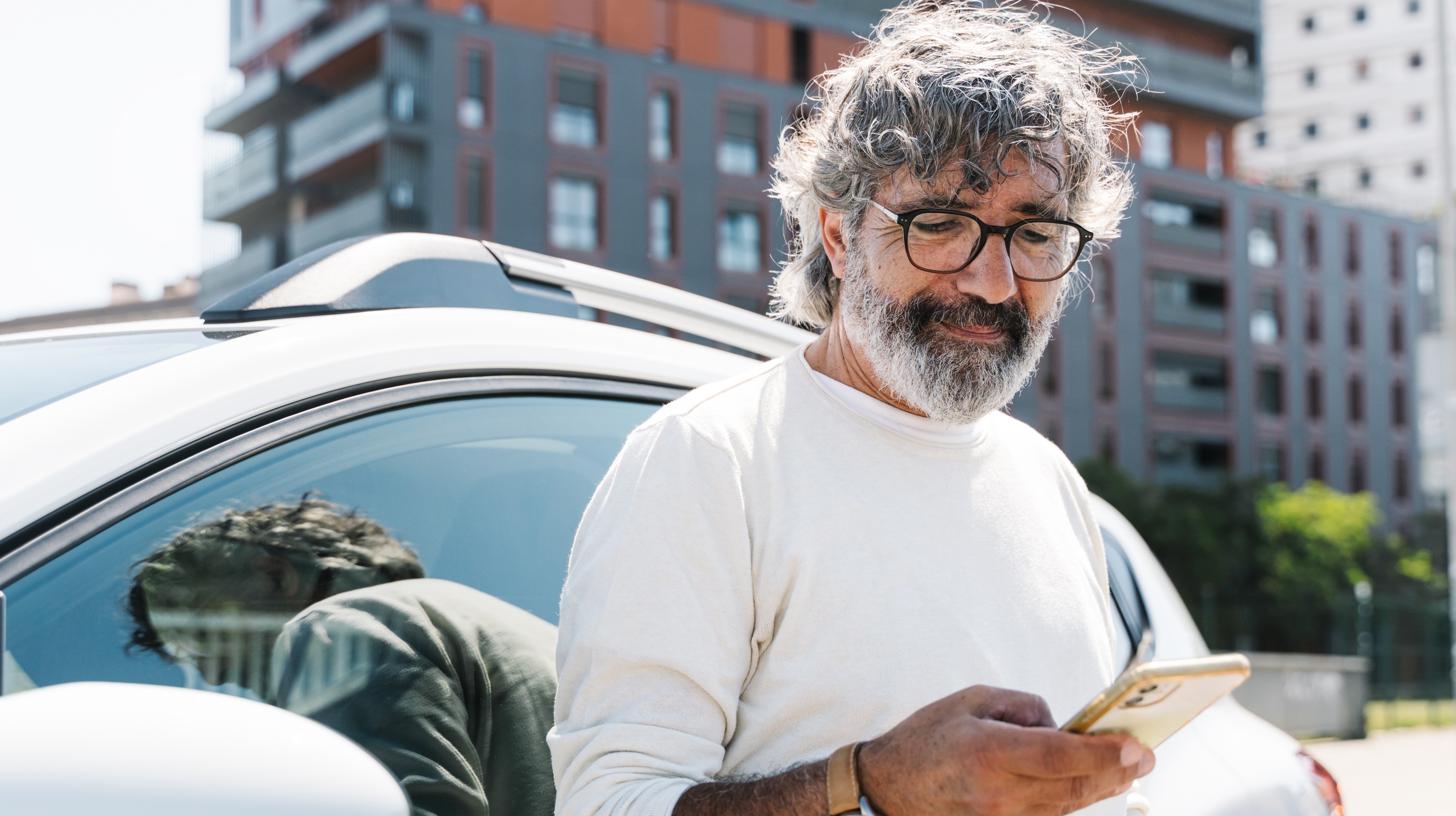 A senior man looks at his phone while leaning against his car.