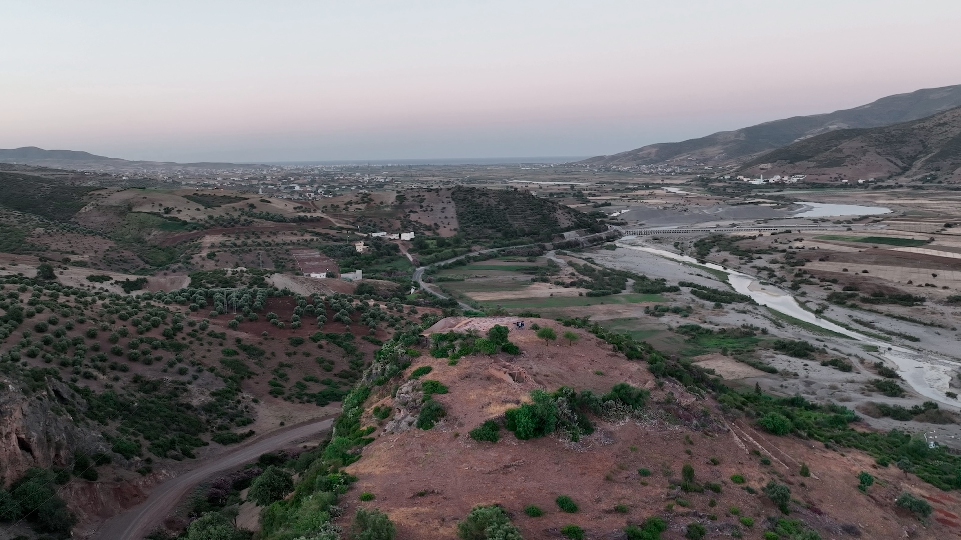 a wide view of an archaeological site iwth hills, scrubby vegetation, a winding stream and towns visible in the distance