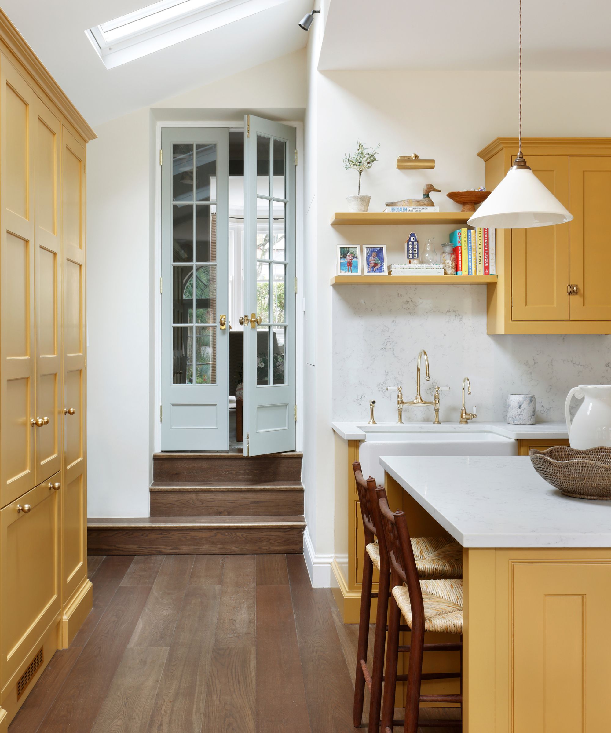 Kitchen with cabinetry painted in India Yellow by Farrow &amp;amp; Ball, wooden flooring, glass pale blue doors, marble countertops and backsplash, and a low white pendant light above the island.