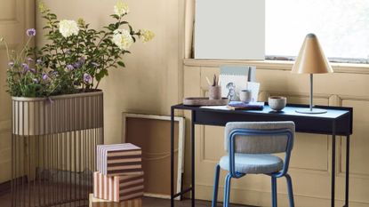 A rustic study with flowers in a standing wire planter near a stack of boxes a turned over canvas, a desk with a lamp and art supplies by a window with a metal chair