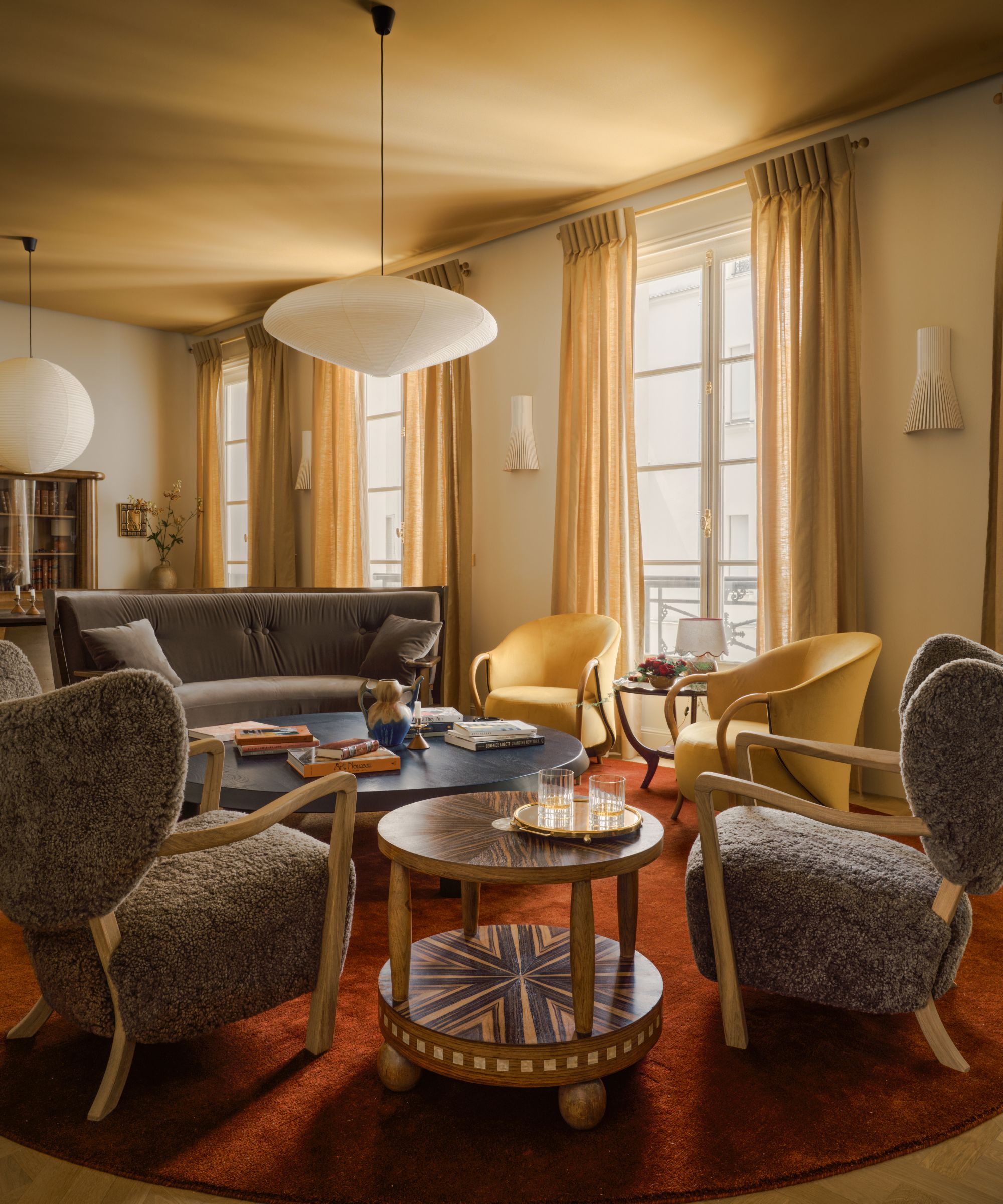 A large living room with an ochre ceiling, off-white walls, ochre curtains on three floor-to-ceiling windows, a red rug, blue and yellow armchairs.