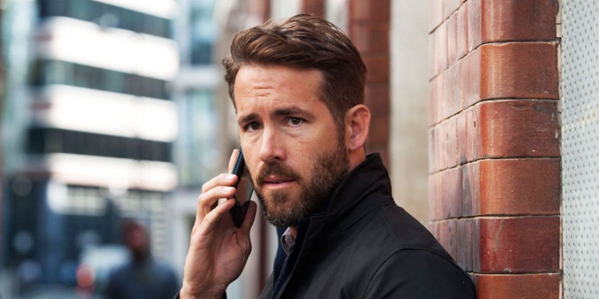 Ryan Reynolds holds a cell phone to his ear while standing against a brick wall.