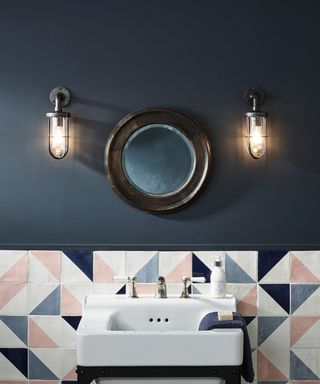 A navy blue bathroom with two silver wall sconces on the wall, a dark brown round mirror, and a white, pink, and blue geometric splashback with a white basin on it