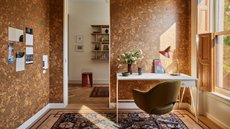 A home office with cork walls, a standing white desk, a green swivel chair, patterned rug, a curved table lamp, and a vase of flowers