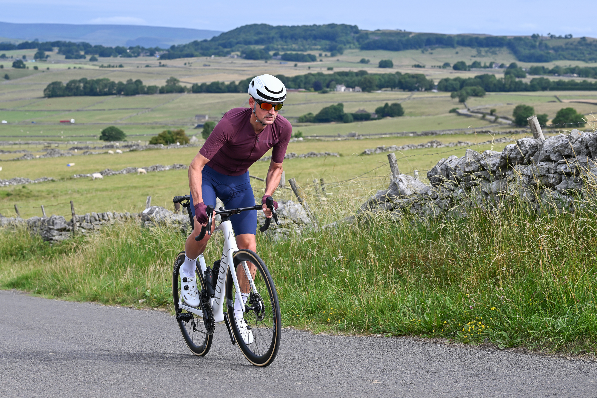 male cyclist wearing a purple jersey, white helmet and sunglasses riding a white bike uphill out of the saddle