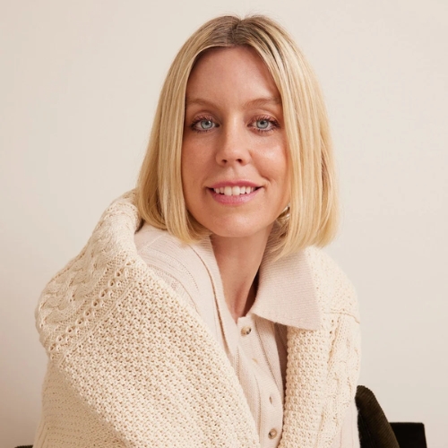 Image of a woman with a blonde bob wearing a cream, knit shirt with a cream, knit sweater wrapped around her, sitting in front of a cream background.
