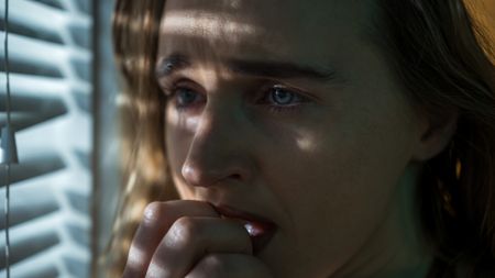 A close up of a woman with wavy brown hair holds her hand to her mouth. She stands next to closed window blinds and looks to the left of the image