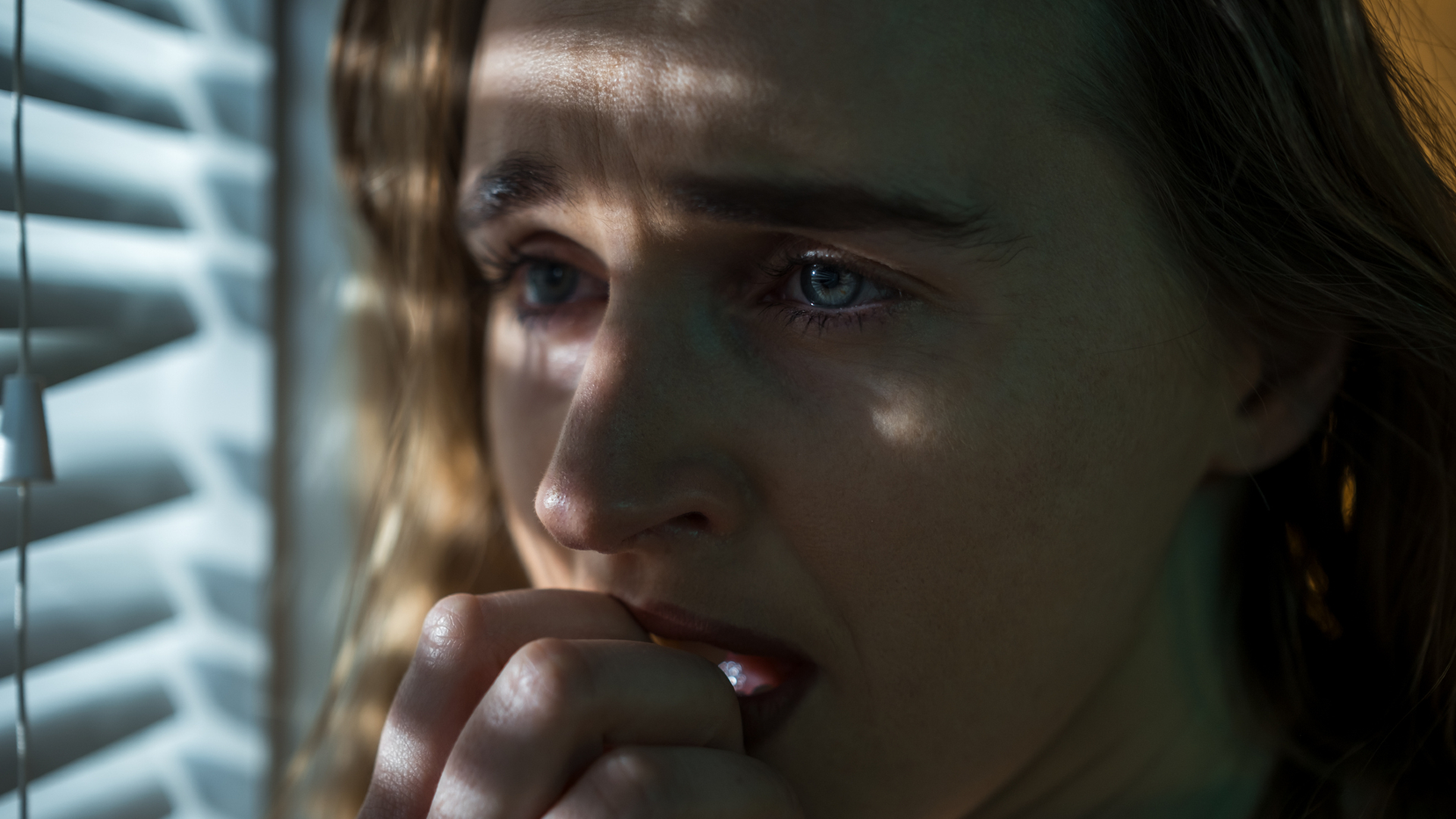A close up of a woman with wavy brown hair holds her hand to her mouth. She stands next to closed window blinds and looks to the left of the image
