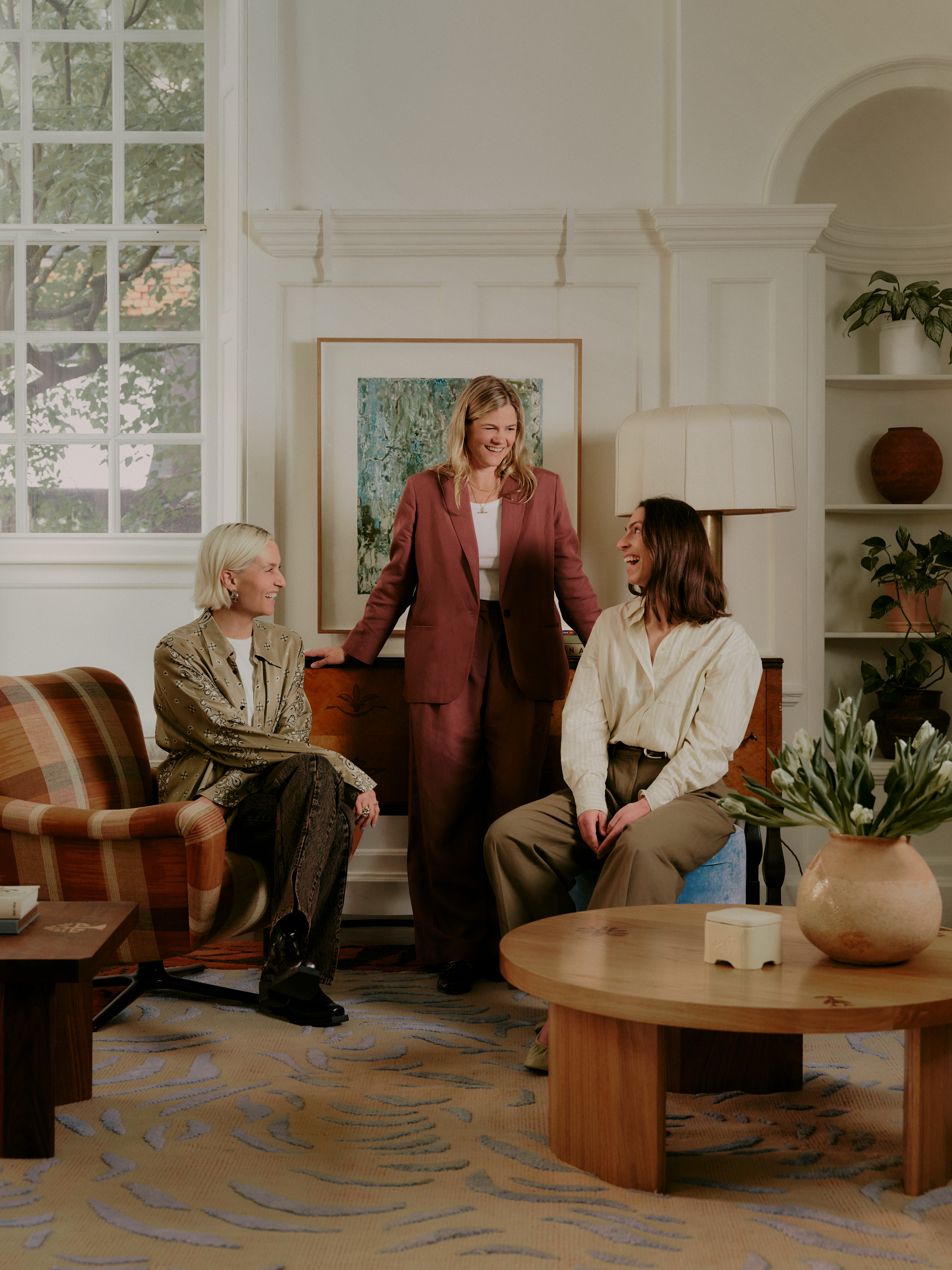 A group of women dressed in oversized suits in beige, pale green, and terracotta sit in a naturally lit room decorated with wooden furniture, plants, and ceramics while smiling at each other.