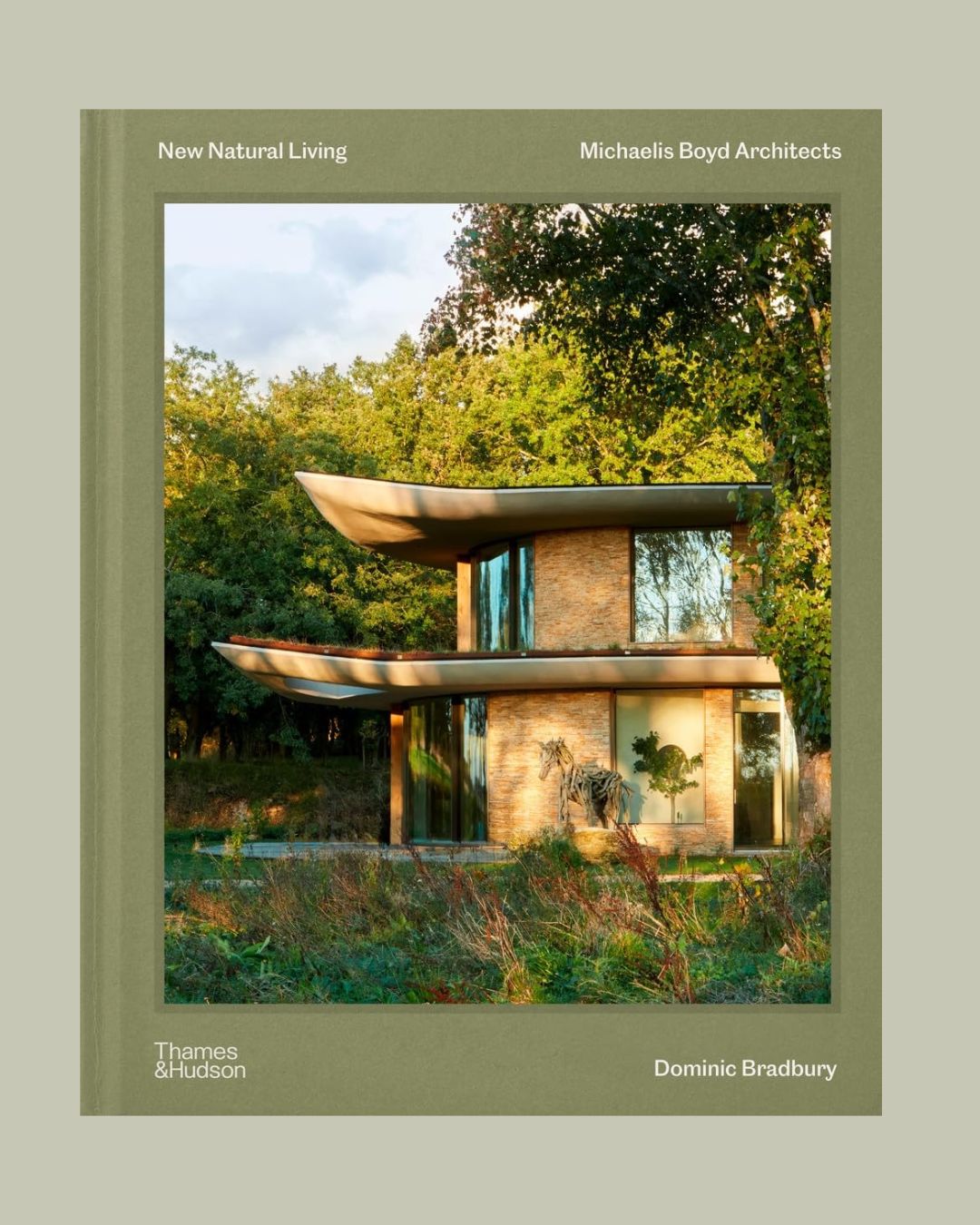 A cover of a coffee table book with a dark green background and the sun-lit facade of a sculpturally built house with two overlapping roofs, wide floor-to-ceiling windows, and greenery all around.
