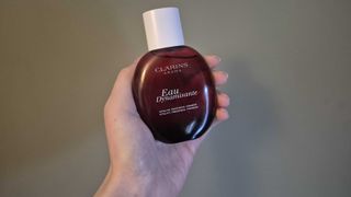 A close-up of Digital Beauty Writer, Naomi Jamieson's hand whilst holding the Clarins Eau Dynamisante perfume fragrance treatment bottle, in front of a dark green backdrop