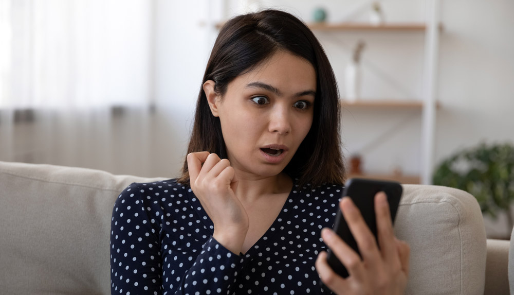 Dark-haired woman looking at smartphone screen in shock.