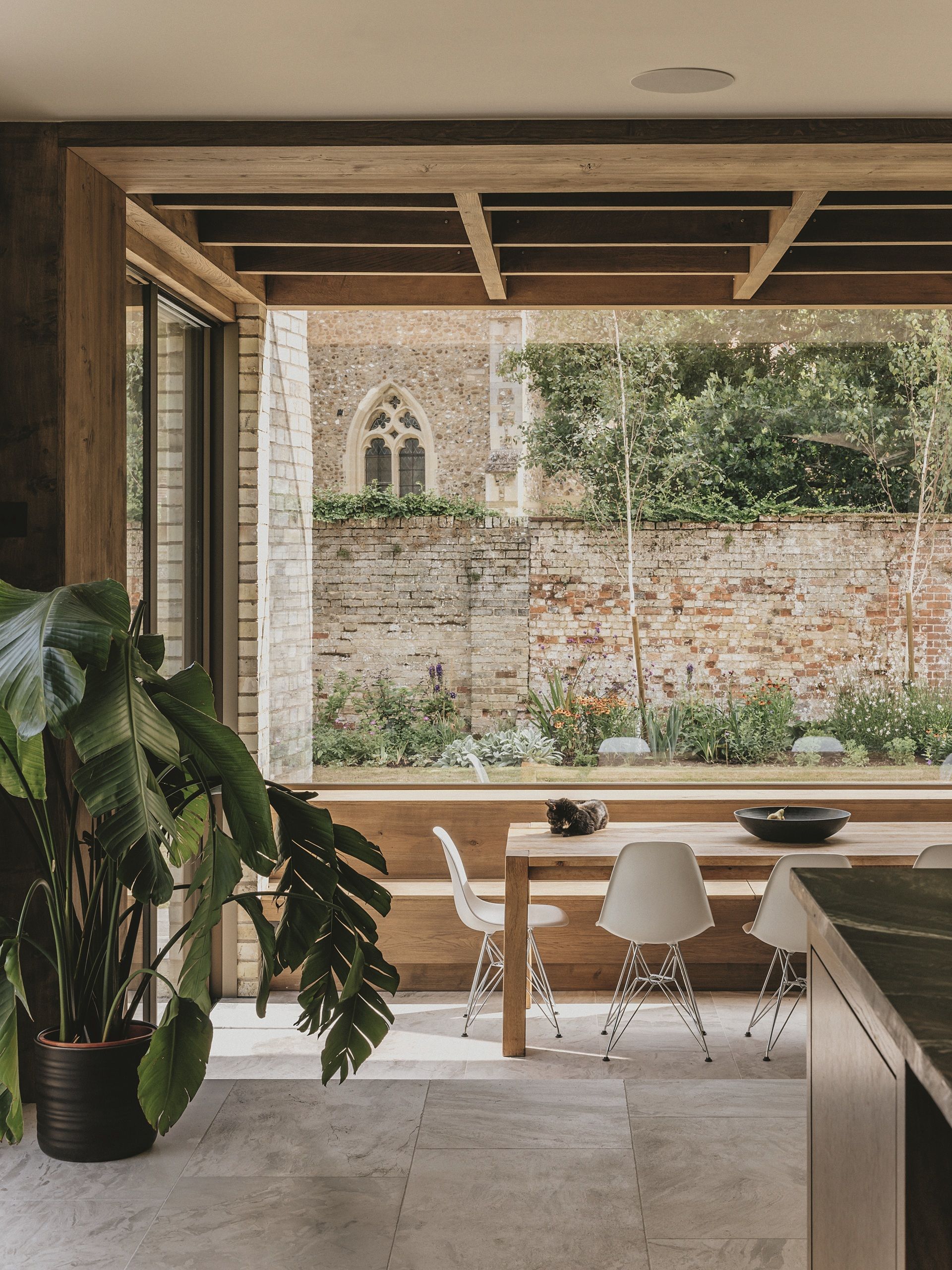 modern extension with exposed timber ceiling, tiled floor, brick walls, large glass window above dining table and timber banquette, with potted monstera