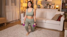 A woman practices lunges at home while holding dumbbells in her hands. 