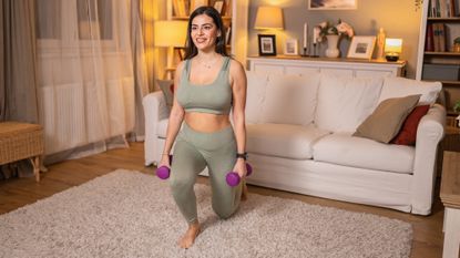A woman practices lunges at home while holding dumbbells in her hands. 