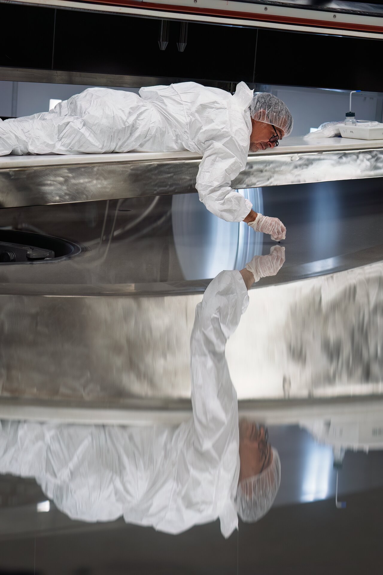 A man wearing a white jumpsuit and hair net and white gloves lays on a glass mirror and polishes it.