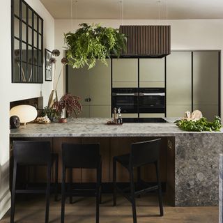 A warm kitchen with matte green cabinets, a stone island, black chairs, and hanging plants