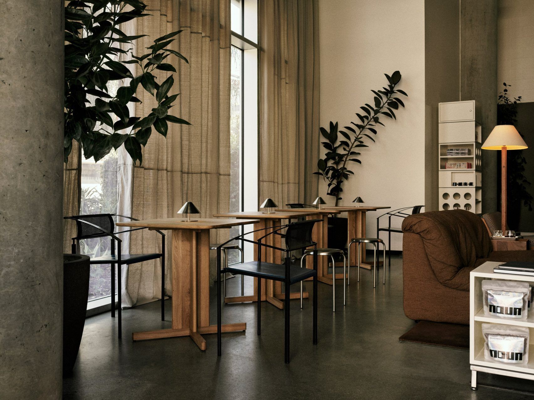 A home-looking coffee shop decorated with modernist furniture including spherical paper lanterns as lights, wood and chrome minimalist furniture, chrome table lamps, and thriving plants.