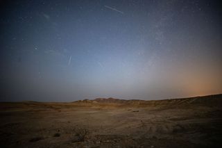 Numerous perseid meteors streak across the star filled sky above a barren desert landscape.