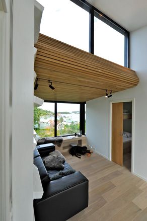 Interior view of a space at Trekronekabin featuring a wood slat covered ceiling, black bar spotlights, white walls, wood flooring, a black sofa and a wooden unit under the windows. There is a cupboard with a partially open wooden door and a dog is lounging on the floor
