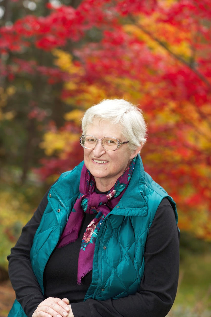 Headshot of woman with short, white hair, glasses, wearing a black top and teal gillet