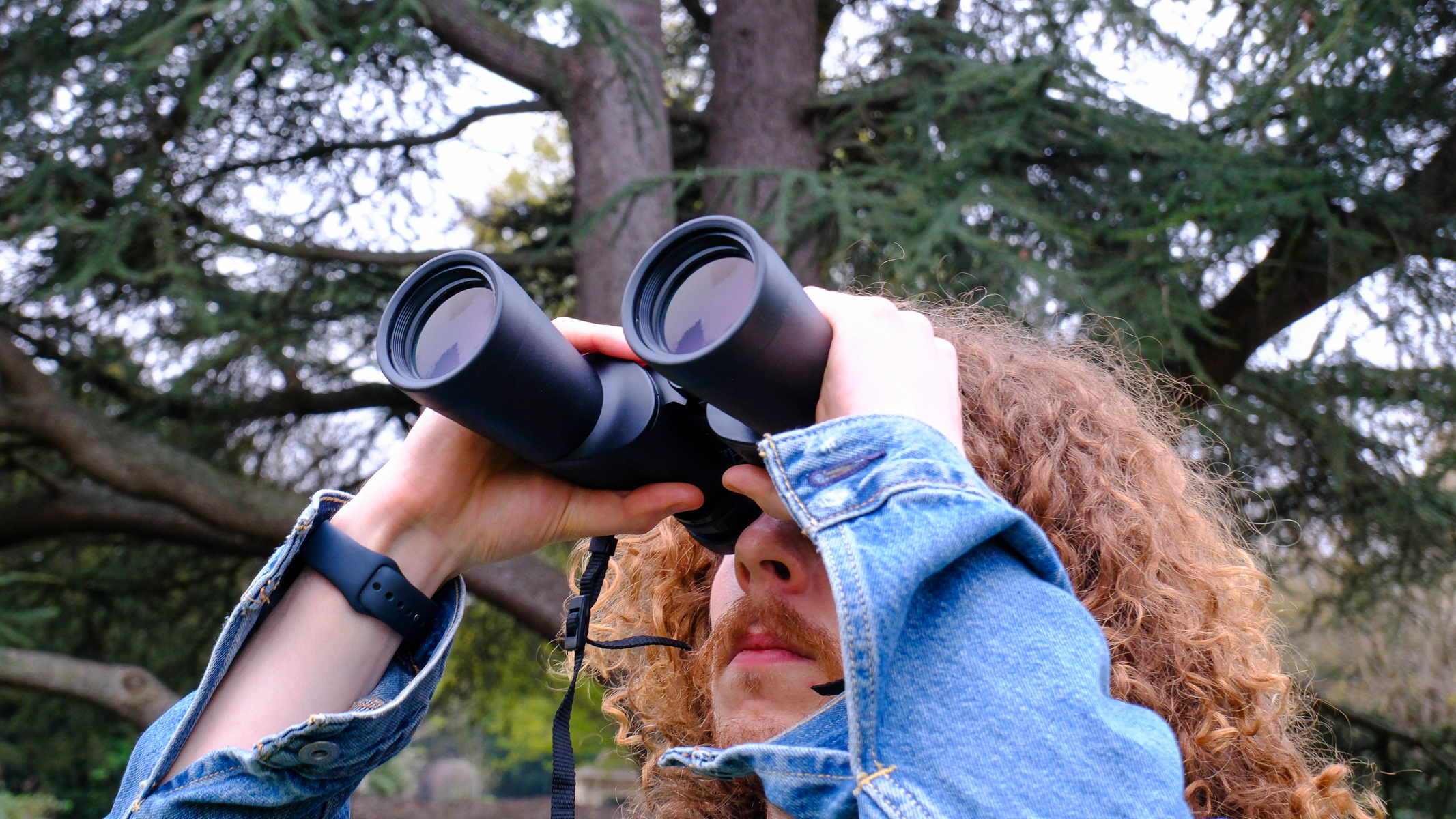 A man with long curly hair looking through the Nikon Action 16x50 binoculars with a tree in the background.