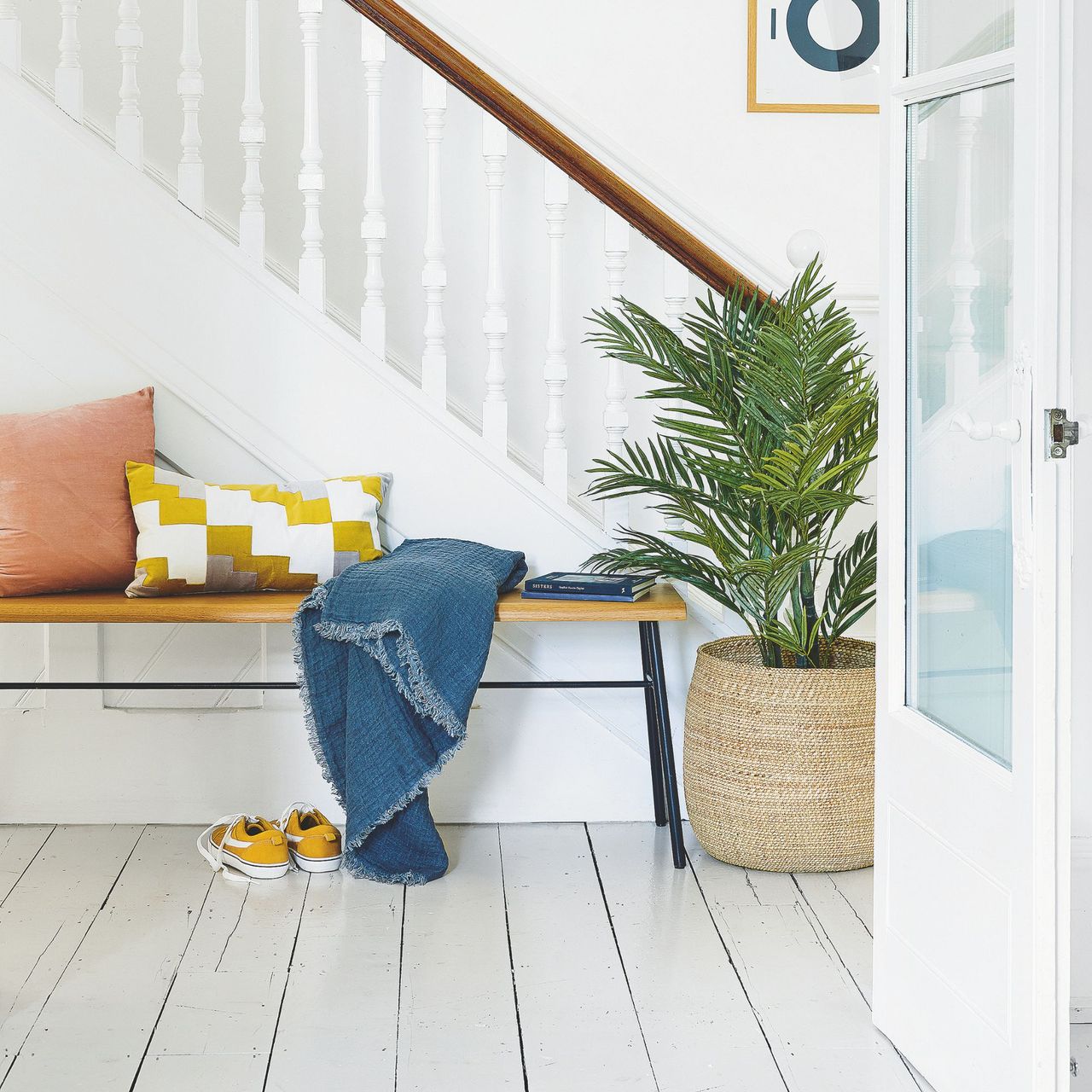 Area under the stairs with white floorboards, white walls and white stair railing, and a wooden bench with cushions on it and a palm plant next to it
